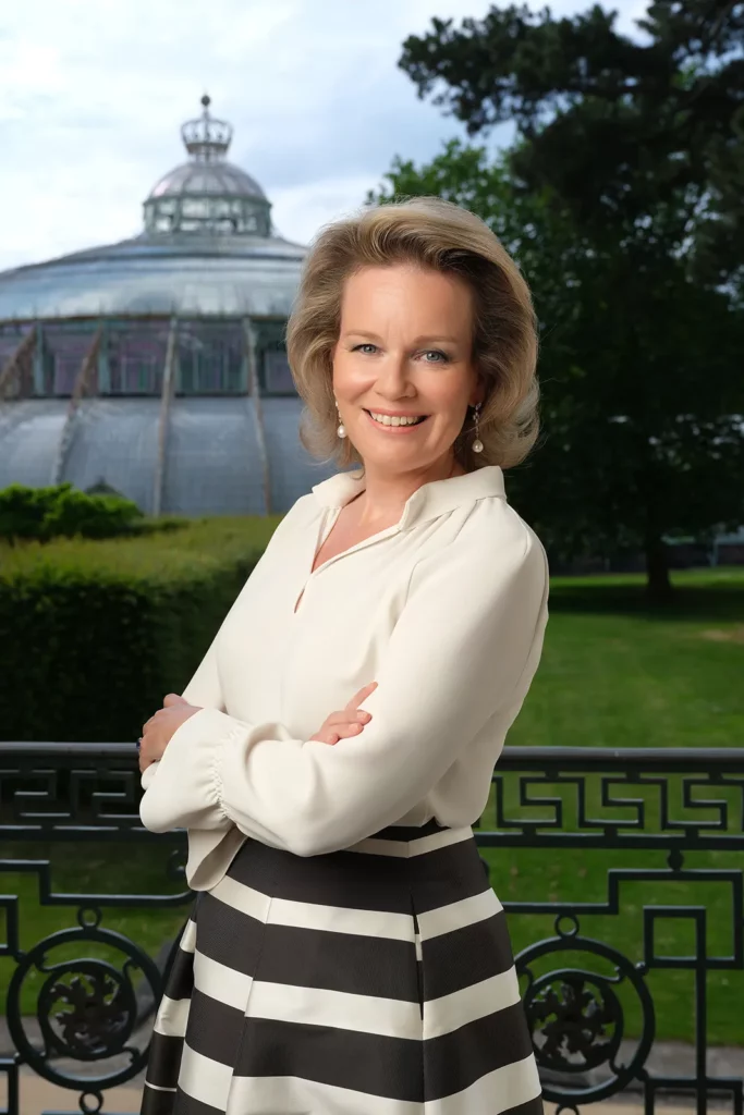 Portrait de la Reine Mathilde debout devant une fenêtre donnant sur les Grandes Serres de Laeken.
