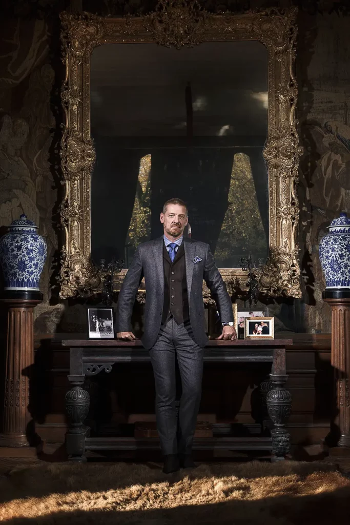 Gérald Watelet en costume gris appuyé sur une commode ancienne entre deux vases de Chine, devant un grand miroir doré.