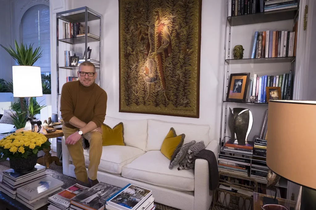Gérald Watelet posant dans son salon bibliothèque avec tapisserie et étagères vintage.