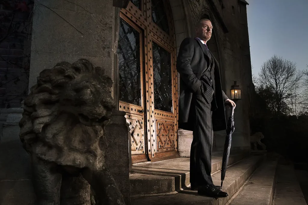Gérald Watelet en costume rayé et manteau noir, appuyé sur un parapluie devant l'entrée d'un château à la tombée de la nuit.
