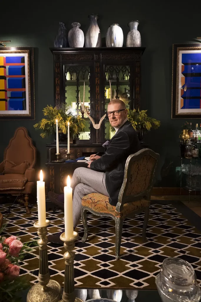 Gérald Watelet assis devant son secrétaire ancien, écrivant entouré de tableaux modernes.