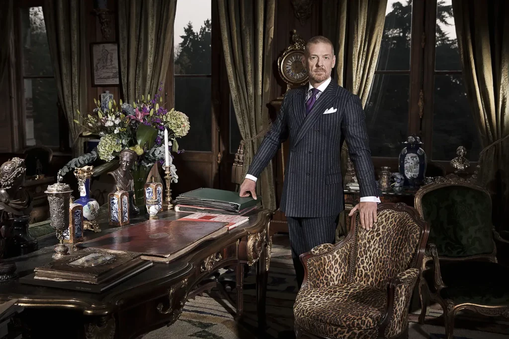 Gérald Watelet debout dans un bureau de style Louis XV richement décoré d'antiquités, une main sur une chaise XIXème en peau de panthère.