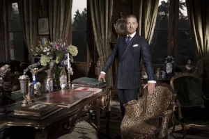 Gérald Watelet debout dans un bureau de style Louis XV richement décoré d'antiquités, une main sur une chaise XIXème en peau de panthère.