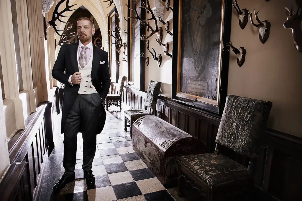 Gérald Watelet en costume queue-de-pie dans un couloir de château avec trophées de chasse, tableaux anciens et sol damier noir et blanc.