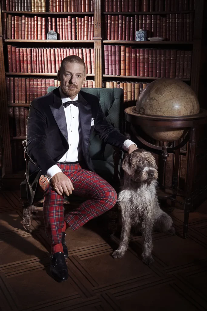 Gérald Watelet en smoking et pantalon tartan assis dans une bibliothèque ancienne avec un chien Griffon Korthals et une mappemonde.