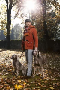 Gérald Watelet en veste de laine Bark et pantalon PT01 avec deux chiens Braque de Weimar en laisse dans un parc en automne.