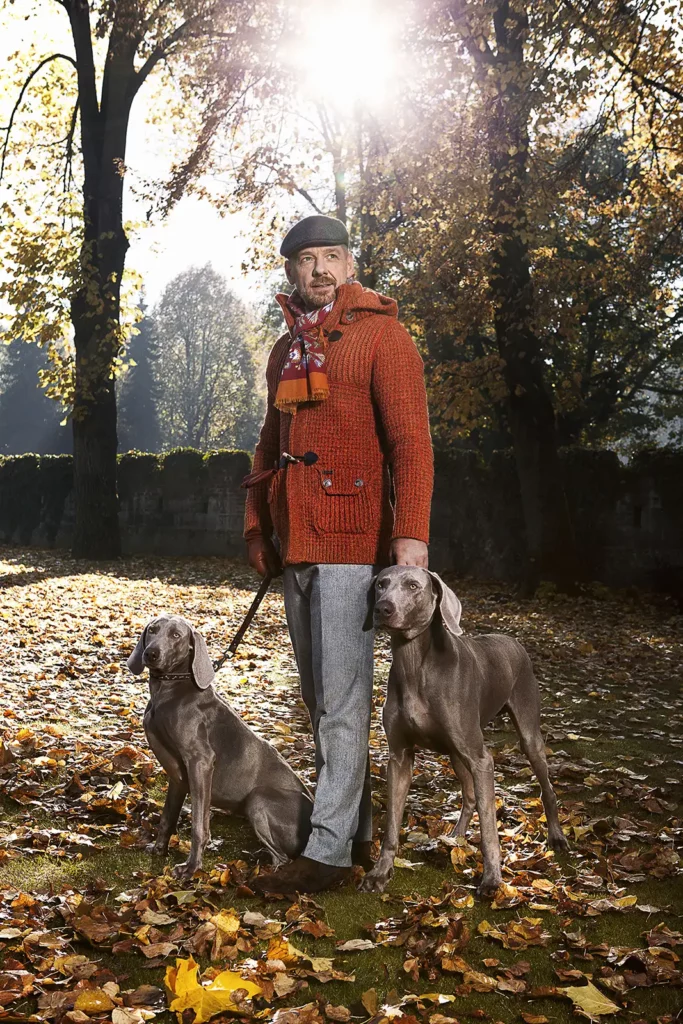 Gérald Watelet en veste de laine Bark et pantalon PT01 avec deux chiens Braque de Weimar en laisse dans un parc en automne.