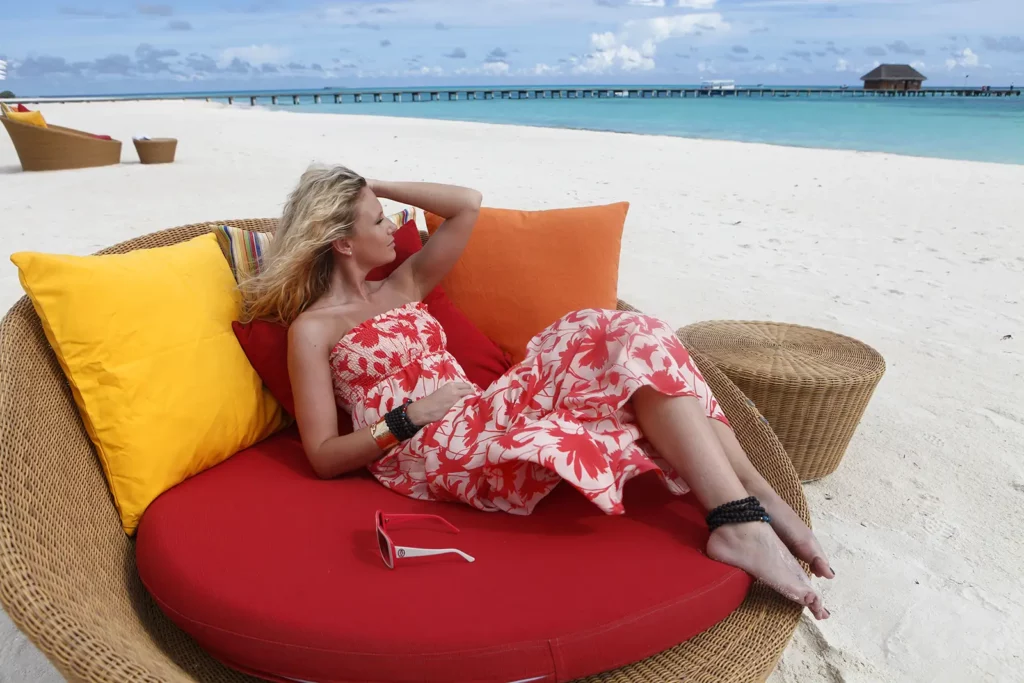 Julie Taton posant dans un grand fauteuil en osier avec coussins colorés sur une plage du Kanuhura aux Maldives.
