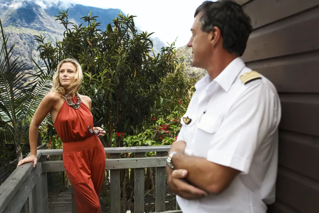 Julie Taton en ensemble pantalon bordeaux sur la terrasse d'une maison créole typique à Mafate, regardant le paysage sous l'œil du pilote d'hélicoptère. Photo : Michel Gronemberger.