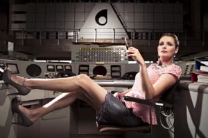 Sandrine Scourneau assise au poste de contrôle des antennes de Wavre avec un porte-cigarette, logo RTB en fond.