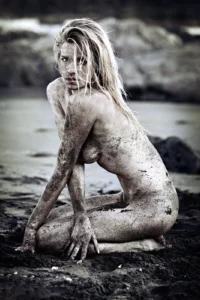Portrait d'art de Sophie Pendeville accroupie sur une plage de sable noir volcanique à la Réunion. Effet de bichromie et fort contraste entre la peau couverte de sable et l'environnement sombre. Photographie artistique de Michel Gronemberger.