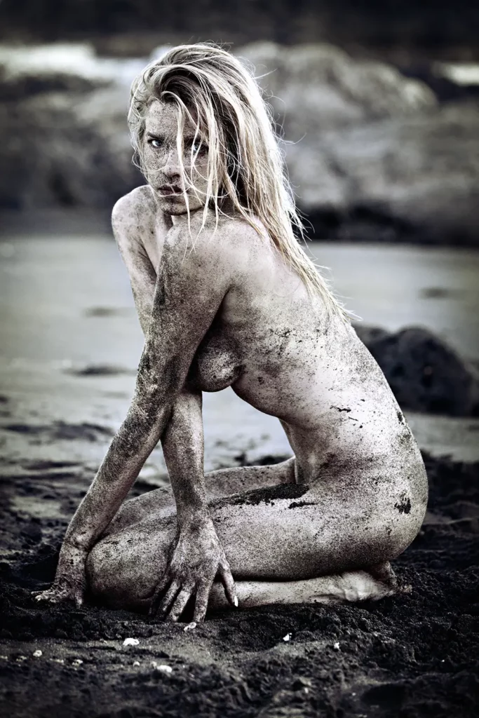 Portrait d'art de Sophie Pendeville accroupie sur une plage de sable noir volcanique à la Réunion. Effet de bichromie et fort contraste entre la peau couverte de sable et l'environnement sombre. Photographie artistique de Michel Gronemberger.