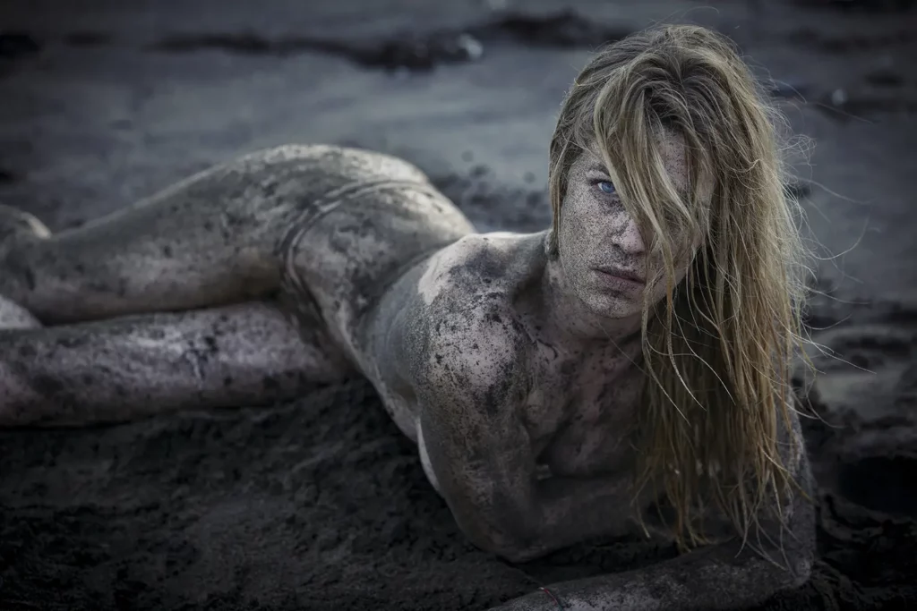 Sophie Pendeville de face 3/4, allongée sur le sable noir de la Réunion, gros plan sur ses yeux bleus intenses. Peau couverte de sable volcanique. Portrait réalisé par le photographe de célébrités Michel Gronemberger.