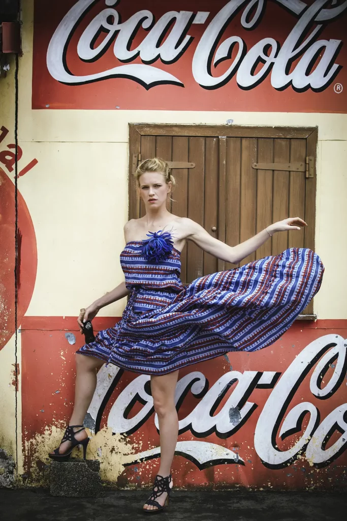 Sophie Pendeville en longue robe volante et talons hauts, tenant une bouteille de Coca-Cola en verre devant la façade jaunie d'un vieux bar vintage à la Réunion. Style photo d'époque. Photo : Michel Gronemberger.