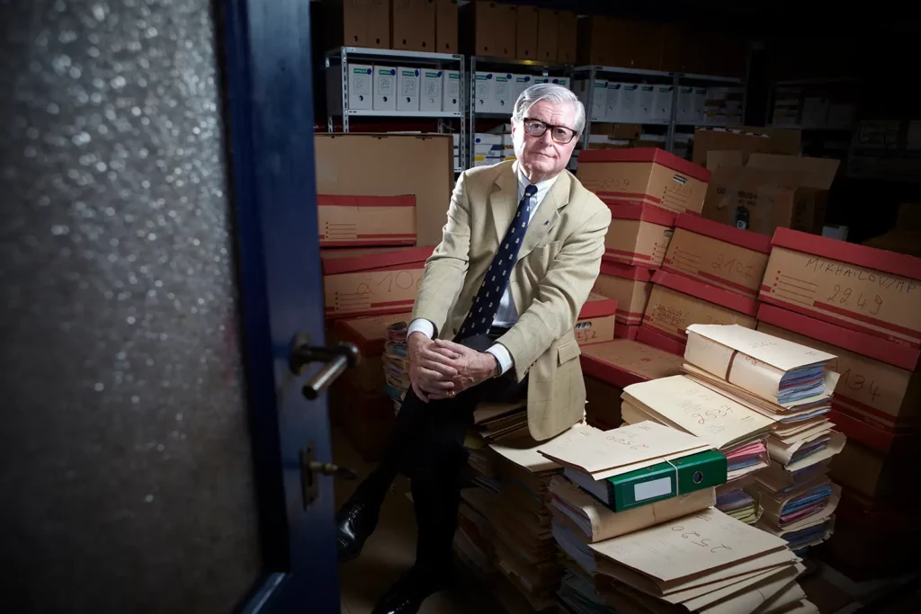 Xavier Magnée assis sur des piles de dossiers dans sa cave, photographie de reportage par Marc Gronemberger.