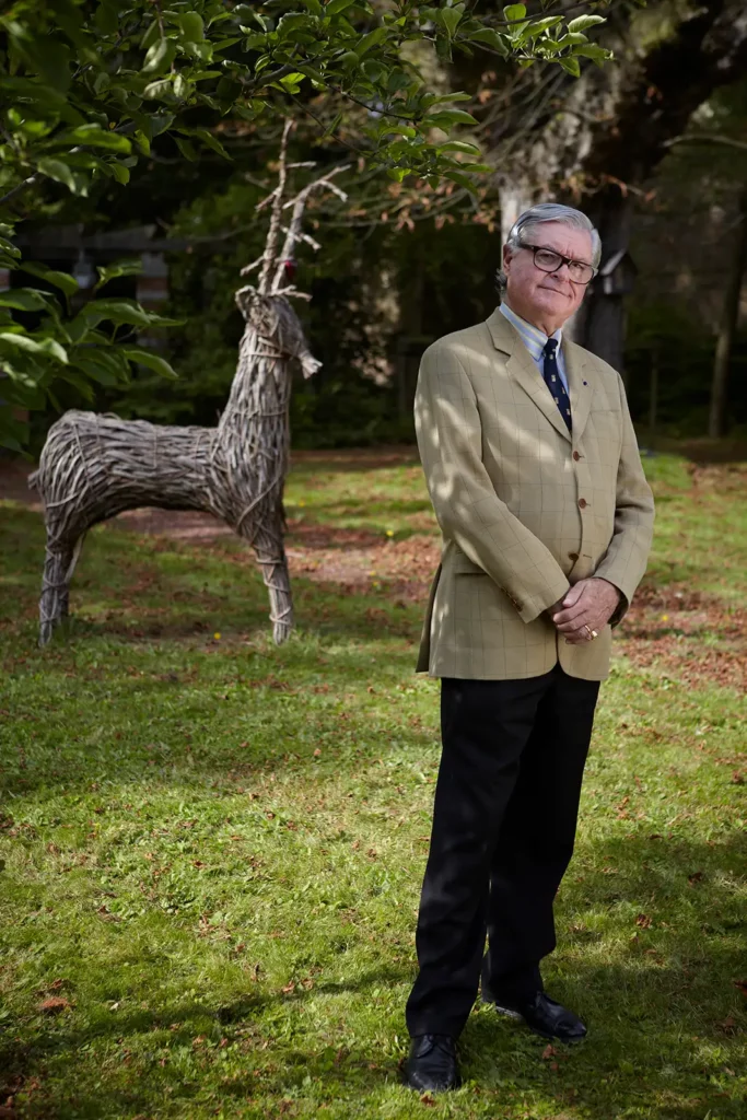 Portrait de Xavier Magnée debout dans un jardin avec une statue de cerf en bois, photographie en extérieur par Marc Gronemberger.