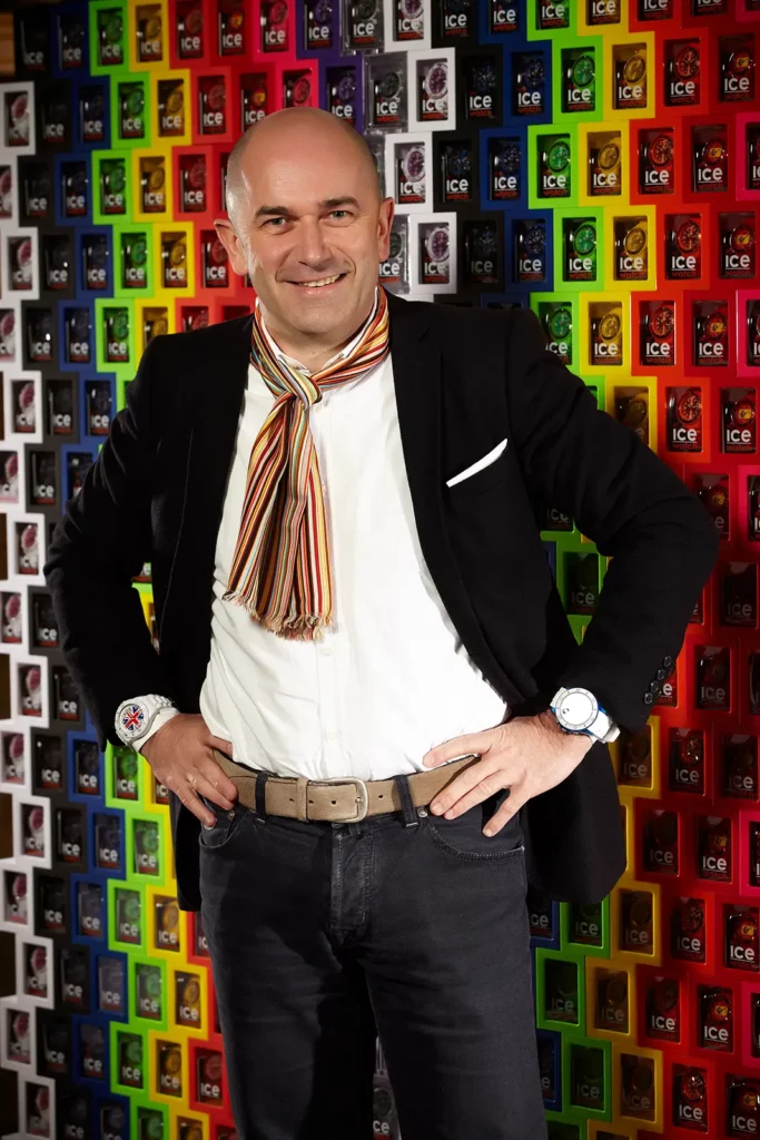 Portrait de Jean-Pierre Lutgen, CEO d'Ice-Watch, mains sur les hanches montrant deux montres devant un mur de boîtes colorées.