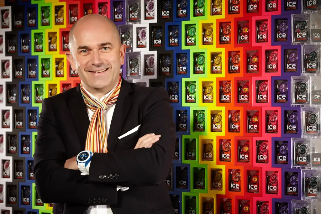 Jean-Pierre Lutgen, CEO d'Ice-Watch, souriant les bras croisés devant un mur de boîtes de montres colorées.