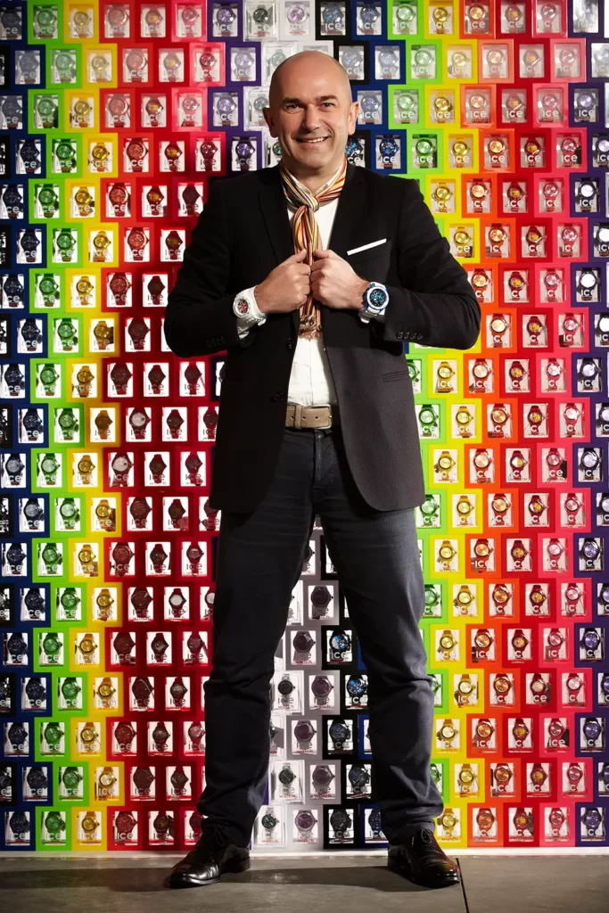 Jean-Pierre Lutgen, CEO d'Ice-Watch, en costume noir avec une montre à chaque poignet devant un mur de boîtes colorées.