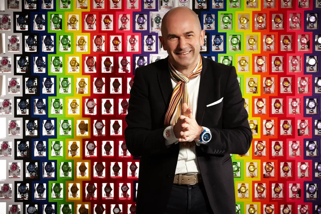 Jean-Pierre Lutgen, CEO d'Ice-Watch, souriant avec les mains jointes devant un mur de boîtes de montres.