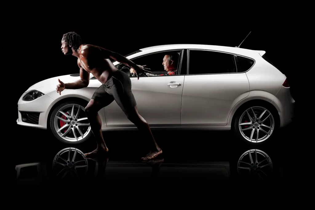 Romelu Lukaku de profil, pieds nus en short, prêt à sprinter contre une voiture de course SEAT pilotée par Marc Duez.