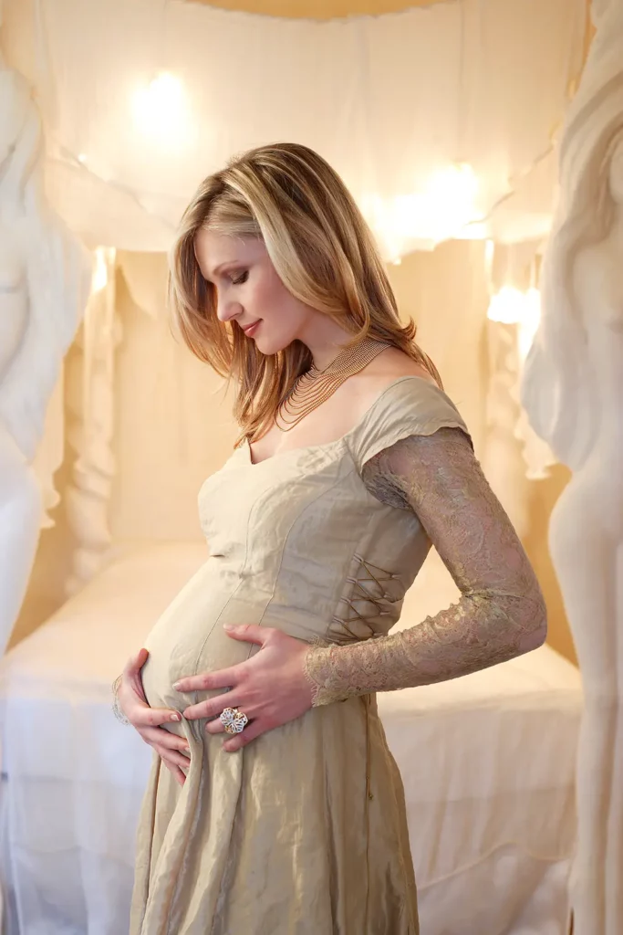 Portrait de profil de Sandrine Corman enceinte regardant son ventre, devant un lit à baldaquin dans la suite Kaat Tilley.