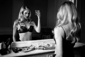 Sophie Pendeville en boudoir chic, soutien-gorge devant un miroir, avec un revolver Colt et un plateau d'argent sur une cheminée.