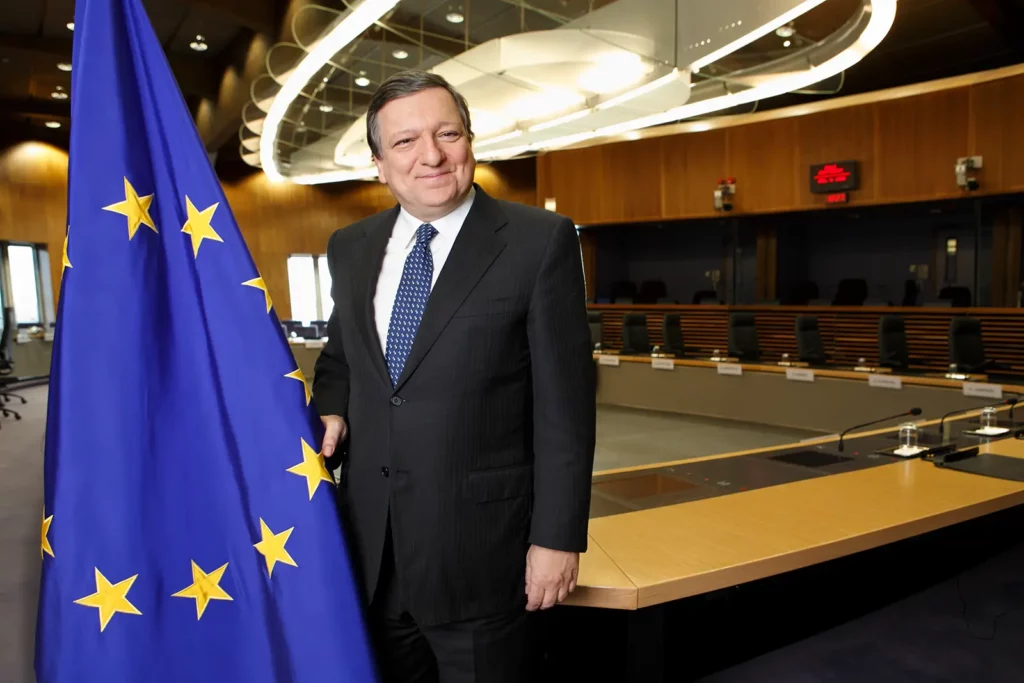 Portrait de José Manuel Barroso debout dans la salle du conseil des chefs d'État, tenant le drapeau européen d'une main avec un sourire bienveillant. Cadrage américain décentré à gauche.