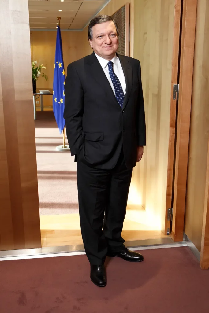 Portrait de plein pied de José Manuel Barroso souriant à l'entrée de son bureau au Berlaymont, avec le drapeau Européen visible en arrière-plan.