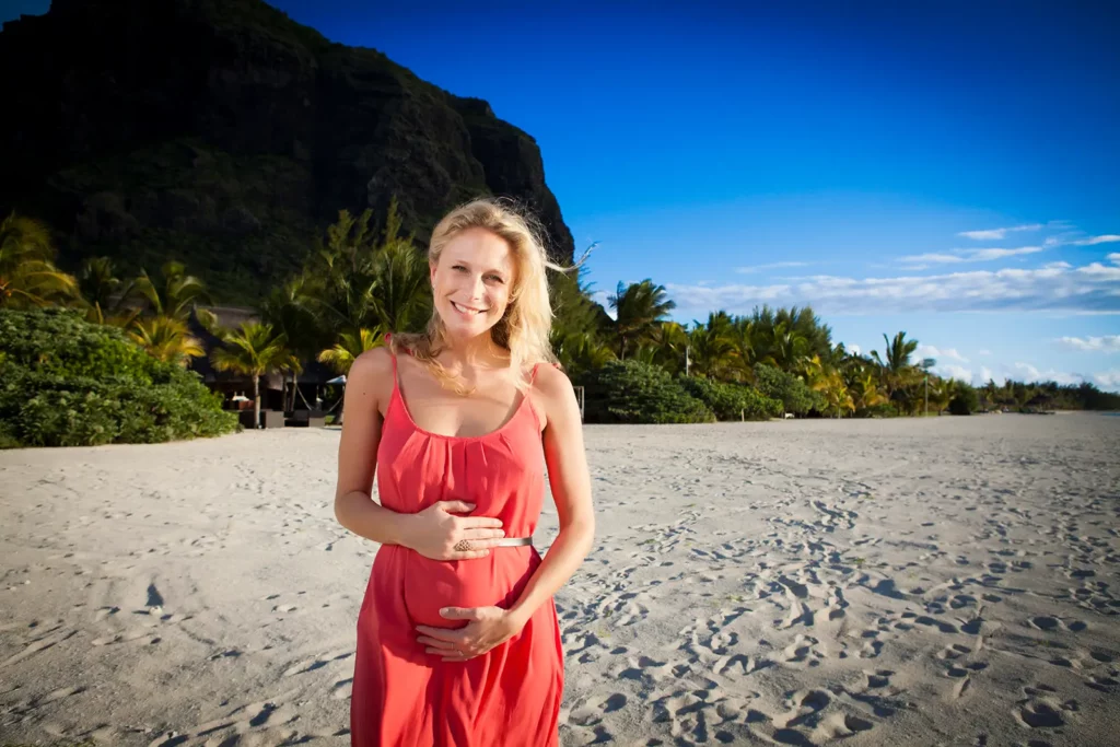 Portrait horizontal de Julie Taton enceinte de 35 semaines, mains sur le ventre et robe cintrée sur la plage du Dinarobin à l'île Maurice avec le Morne en arrière-plan.
