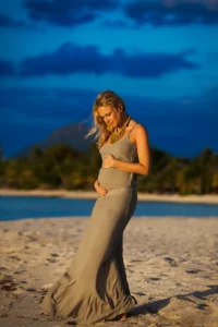 Portrait vertical de Julie Taton enceinte à l'île Maurice, sa longue robe kaki volant au vent au coucher du soleil.