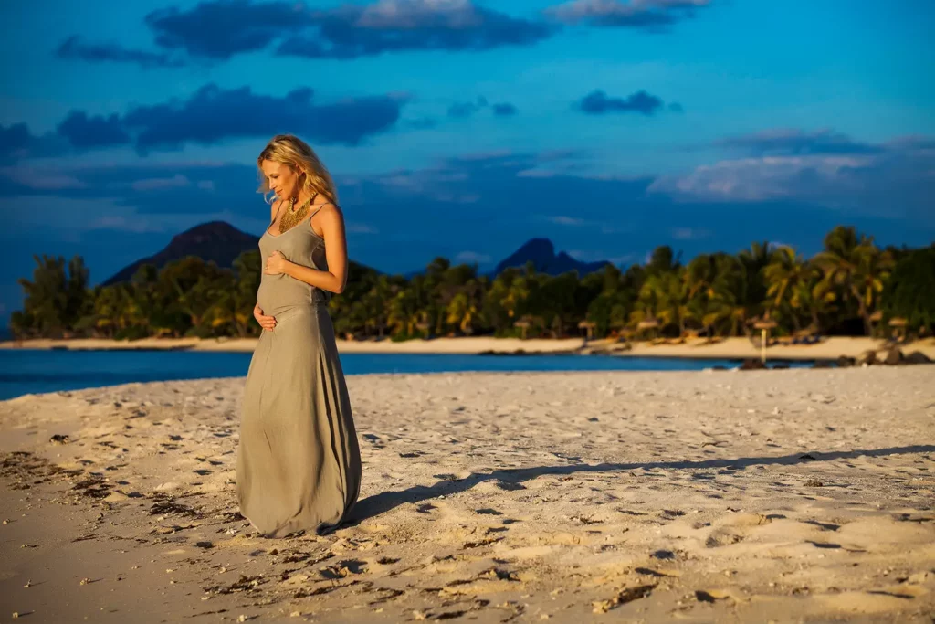 Silhouette complète de Julie Taton enceinte en longue robe kaki et collier doré, les deux mains sur le ventre au coucher du soleil à l'île Maurice.