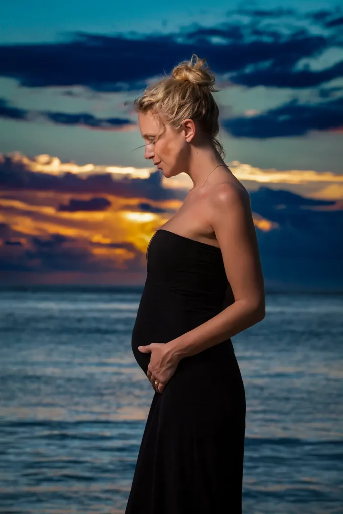 Portrait de profil de Julie Taton enceinte en robe bustier noire sur la plage de l'île Maurice, regardant son ventre au coucher du soleil voilé.