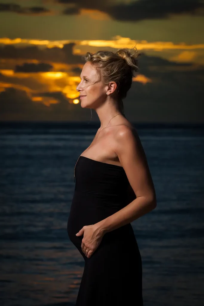 Portrait de profil complet de Julie Taton enceinte en robe bustier noire à l'île Maurice, regardant l'horizon avec un éclairage flash subtil.
