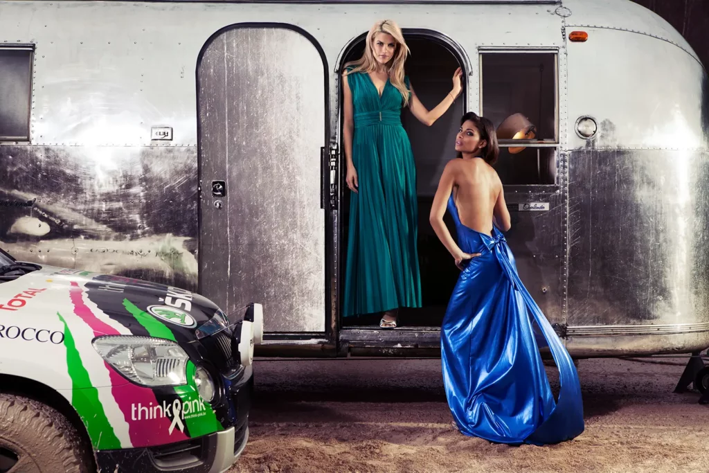 Tanja Dexters et Tatiana Silva posant devant l'imposante caravane Airstream et la Skoda préparée pour le Rallye des Gazelles. Tanja est dans l'entrée en robe verte, Tatiana pieds au sol en robe bleue dos nu.