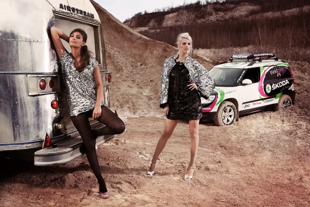 Portrait de Tatiana Silva en robe argentée et bas nylon, appuyée à une caravane Airstream aux côtés de Tanja Dexters. En arrière-plan, la Skoda du Rallye des Gazelles.