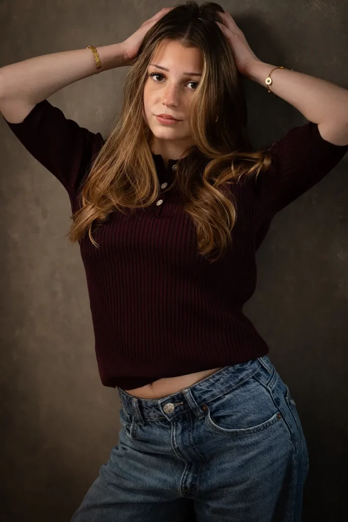 Photographe book modèle Bruxelles, jeune fille en top bordeaux posant debout les mains dans les cheveux devant le fond gris.