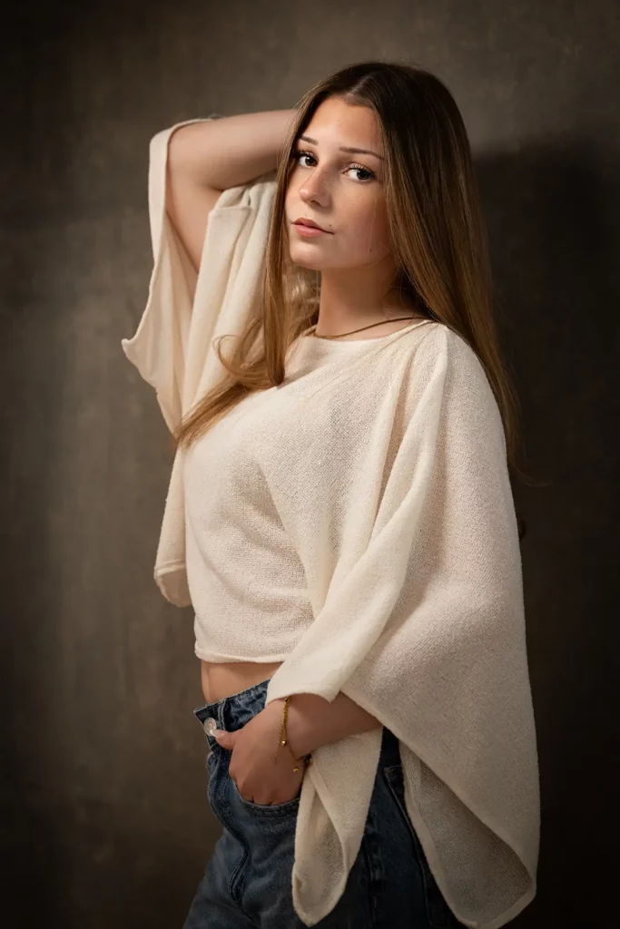 Photographe book modèle Bruxelles, jeune femme de profil en top blanc crème à manches amples, le coude appuyé contre le fond.
