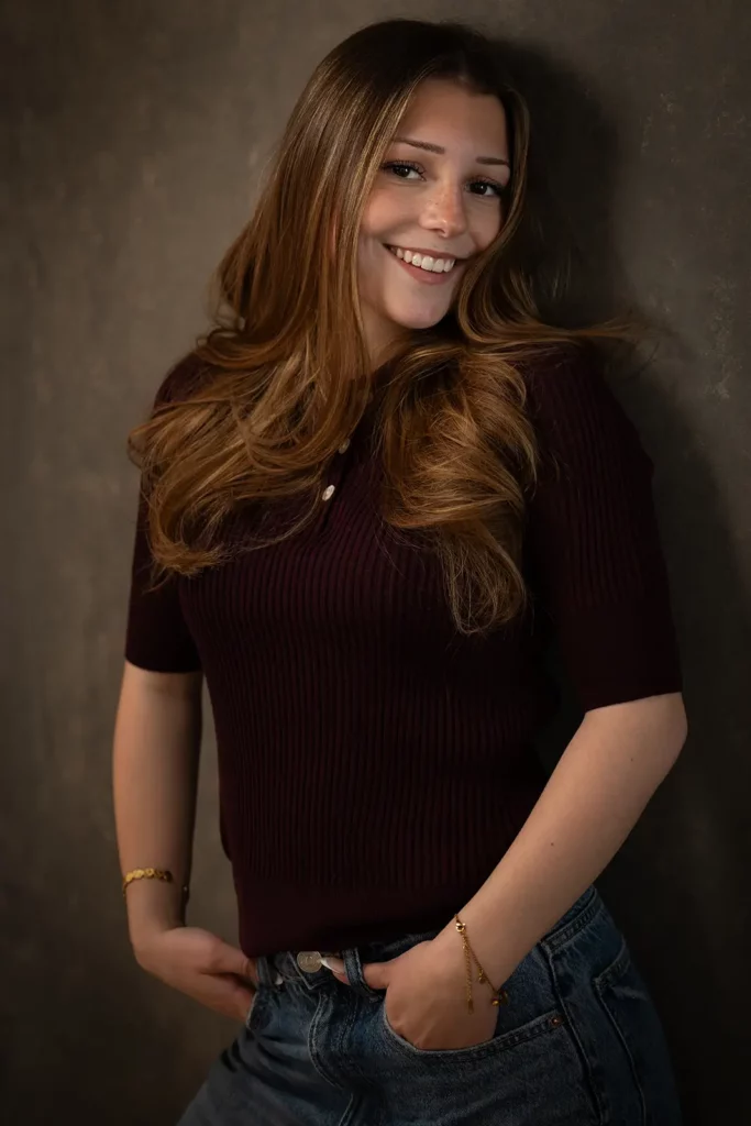 Photographe book modèle Bruxelles, jeune femme de 18 ans très souriante en top bordeaux et jeans posant de trois-quarts.