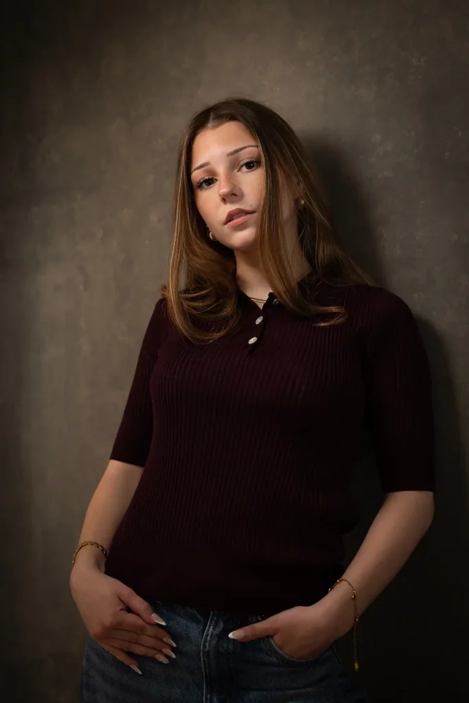Photographe professionnel book modèle Bruxelles, jeune femme en top bordeaux posant de trois-quarts appuyée contre le fond de studio gris.