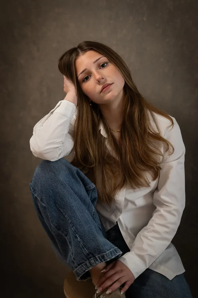 Photographe professionnel book photo à Bruxelles, jeune fille assise sur un tabouret la main contre la tête en chemisier blanc.