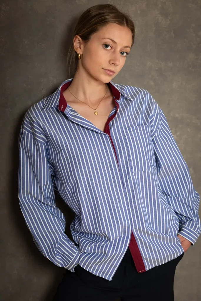 Plan américain debout avec mains dans les poches d'une étudiante pour son portrait professionnel CV, portant une chemise rayée bleue et blanche.