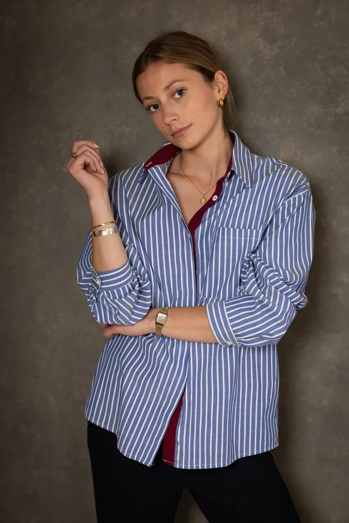 Plan américain d'une jeune femme debout devant un fond gris marbré pour son portrait professionnel CV, portant une chemise rayée bleue et blanche.